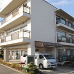3階建ての介護施設の建物外観。白とグレーの外壁、駐車スペースあり。