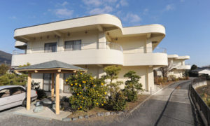 モダンな2階建ての介護施設の建物外観。白を基調とした流線形の設計で、入口前には車寄せスペースがある。